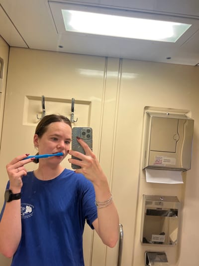 A young woman with brown hair pulled back is brushing her teeth with a blue toothbrush in a well-lit, cream-colored bathroom. She is wearing a blue t-shirt and a watch on her left wrist. She is holding a smartphone in her right hand, which is angled to take a selfie in the mirror. To her right, there is a stainless steel dispenser that appears to be for paper towels, with a roll of white paper hanging out. Below that is another stainless steel dispenser. The bathroom walls are paneled and there are two metal hooks on the wall behind the woman. The lighting comes from a rectangular fluorescent light fixture in the ceiling. The image appears to have been taken inside a train car, as suggested by the interior design and limited space.