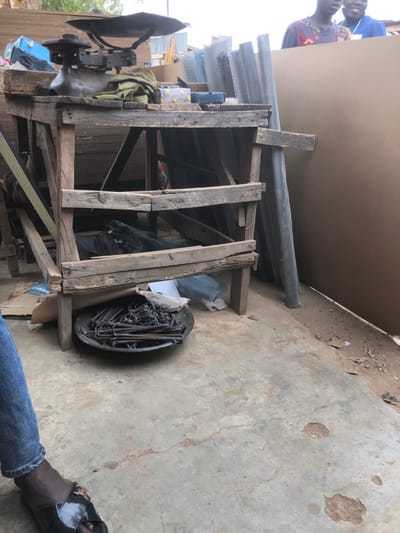 A wooden structure supports a large sheet of material, possibly drywall, with pipes and wire mesh piled on top. Tools and equipment are stored within the structure, including what appears to be a metalworking tool with a large disc. A bucket filled with metal rods sits nearby. The scene unfolds on a weathered concrete surface, with a person wearing jeans and sandals visible in the lower right corner.