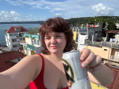 A fair-skinned woman with dark hair and a red top holds up a silver can of beer. She looks toward the camera. Behind her, a hillside and water are seen, and then clouds with a blue sky. To the left of her face, there are buildings with clotheslines and laundry. Buildings with red roofs are seen at the top right. The image is rotated 180 degrees.