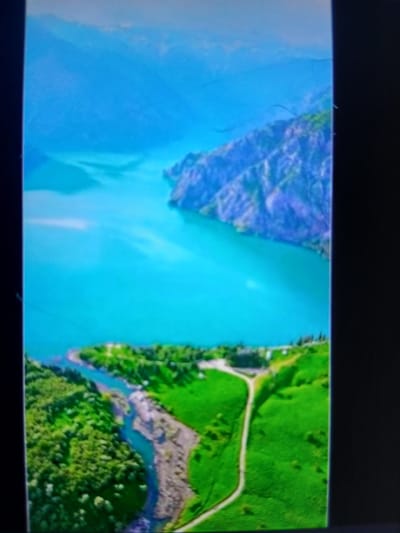 This image depicts a breathtaking aerial view of a mountainous coastal landscape. A vast expanse of turquoise water dominates the scene, meeting rugged, purple-hued mountains in the distance. In the foreground, lush green hills roll down to the water's edge, crisscrossed by a winding dirt road. A river snakes its way through the green terrain, leading to the larger body of water. The scene is tranquil and appears to be a natural landscape with no signs of human habitation or activity, though the road suggests it may be accessible. The location is identified as Auk, Kyrgyzstan.