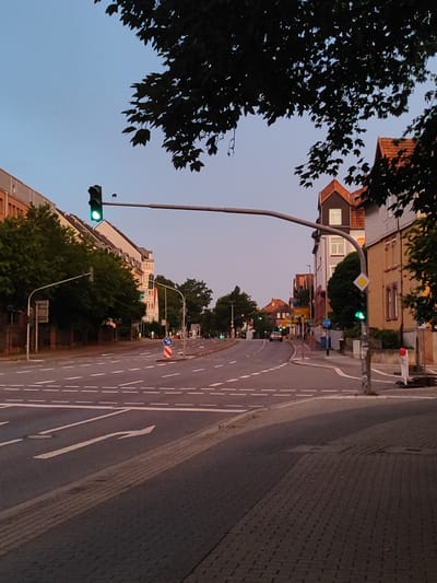 This image depicts a street scene in Göttingen, Germany, likely near an intersection. A traffic light with a green signal is prominently displayed. On the left, a row of apartment buildings with trees in front lines the street. On the right, more buildings and trees are visible, along with another traffic light and a diamond-shaped yellow yield sign. The road is marked with white dashed lines, indicating lanes, and a left-turn arrow is painted on the asphalt in the foreground. The sky is a pale blue, suggesting either early morning or late evening. The overall impression is one of a quiet urban environment during twilight.