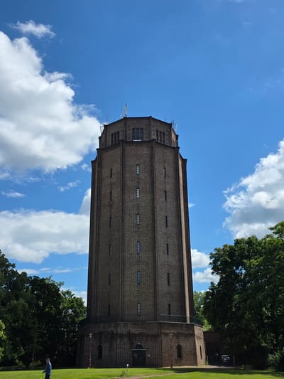 The image shows the water tower in Halle (Saale), Germany. The water tower is a tall, brick structure with a polygonal top. It is surrounded by green trees. The sky is blue with some white clouds. In the foreground, a man in a blue jacket and cap is walking on the grass. There is also a dark-colored car parked near the entrance to the water tower. The water tower is located in a park-like setting with a well-maintained lawn. The overall scene suggests a sunny day in a public space.