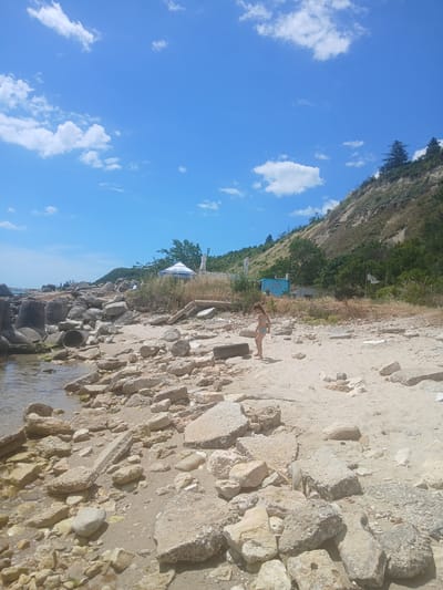 The image depicts a beach scene with a person in the foreground. The person, appearing to be a woman in a bikini, is lying on the sandy shore. The beach is characterized by numerous rocks and stones scattered across the sand, with a body of water visible at the bottom of the frame.

In the background, a steep, vegetated hillside rises up, with trees and dry grass covering its slopes. A small, light blue structure, possibly a building or a food stand, is visible amongst the vegetation. There is also a white umbrella with blue accents, and a flag on a pole. The sky above is a clear blue with scattered white clouds, suggesting a sunny day.

The perspective is from a low angle, looking upwards and across the beach. The lighting indicates it is daytime, likely mid-day or early afternoon, given the bright sunlight and shadows cast by the rocks. The overall impression is one of a relaxed, outdoor setting, likely a remote or less developed beach location. There is some text visible on the blue structure, which reads "CAFE". The text on the umbrella is partially visible and appears to be a brand name.