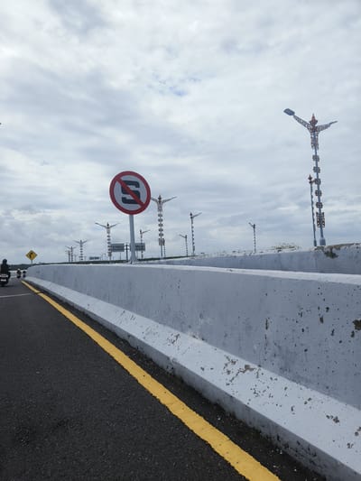 This is an outdoor, eye-level, medium shot of a road with a concrete barrier on the right. The road is paved with asphalt and has a yellow line marking the edge. A few motorcycles are visible on the road. A large, circular sign with a red border and a diagonal red line through the letter "S" is mounted on a pole in the background. There are streetlights with decorative elements in the distance. The sky is overcast with clouds. The scene is located in South Denpasar, Indonesia.