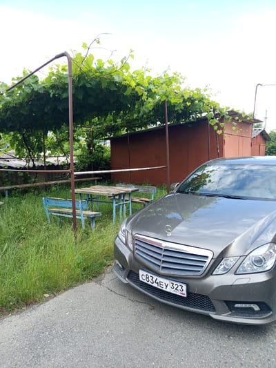 In the city of Otradnaya, Russia, a grey Mercedes-Benz E-Class sedan is parked on an asphalt road adjacent to a grassy area. The car's front is prominently displayed, showing its grille, headlights, and license plate which reads "C834EY 323 RUS". Behind the car, a rustic outdoor seating area is visible, featuring a wooden table and blue benches situated under a vine-covered arbor. To the right of the seating area, there is a weathered, reddish-brown shed or outbuilding. The environment appears to be a rural or semi-rural setting, with overgrown grass and a cloudy sky overhead.