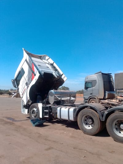 In Cáceres, Brazil, a white semi-truck with its cab tilted upward is parked on a dirt and asphalt lot under a clear blue sky. The truck is undergoing maintenance, with its engine compartment exposed. A toolbox is visible on the ground near the front tire. A silver semi-truck is parked behind it, facing the same direction. In the background, there are trees and a glimpse of other parked vehicles and stacks of what appear to be goods. The ground is dry and dusty.