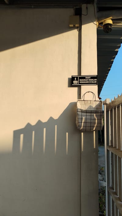 In Banjarsari, Indonesia, a security camera is mounted on a pale yellow building exterior, next to a junction box. A rectangular sign with white text on a black background reads "PAKET MASUKAN KEDALAM KERANJANG DI DALAM" and features an icon of a basket. Below the sign, a fabric basket with a black grid pattern hangs from a hook. The sunlight casts long shadows of vertical elements onto the wall, creating a pattern of elongated rectangles and peaks. A metal gate with vertical bars is visible to the right, along with some greenery. The sky is clear and blue.