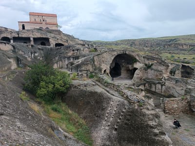 The image depicts the ancient cave city of Uplistsikhe, Georgia. In the foreground, a person wearing a black jacket and gray pants is crouching on a paved area near stone structures. The city is carved into a rocky cliff face, with numerous caves and dwellings visible. A red-tiled roof church, the Uplistsikhe Church, stands prominently atop the cliff. The landscape surrounding the city is characterized by rolling hills covered in dry grass and eroded rock formations. The sky is overcast with gray clouds.
