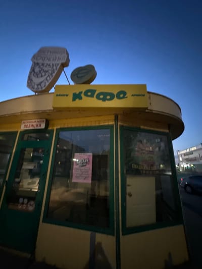 A low-angle, wide-shot photograph captures a small, yellow kiosk with green window frames against a deep blue twilight sky. The kiosk features a prominent yellow sign with green lettering that reads "кафе" (cafe). Above this sign, two illuminated signs are visible: one shaped like a large coffee cup with intricate detailing and text, and a smaller, rounder sign. On the left side of the kiosk, a red and white sign reads "ЭКСТРЕННЫЙ ВЫЗОВ ПОЛИЦИИ" (Emergency Police Call). Inside the kiosk, seen through the windows, are reflections of the surroundings and some indistinct interior elements. A pink flyer is taped to the inside of one window. In the background, a blurred sign for "АТЛАНТ-ФИЛЬМ" (Atlant-Film) is visible along with a street and a moving car. The scene appears to be set in a public urban area at dusk, with the signs suggesting a commercial establishment.