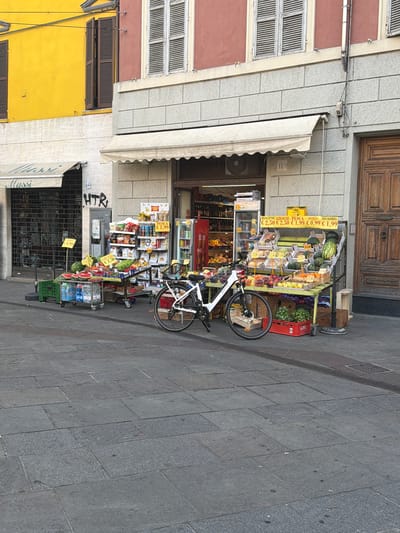 This is an outdoor, eye-level, medium shot of a fruit and vegetable stand in front of a building in Parma, Italy. The stand is laden with a variety of fresh produce, including watermelons, peaches, and possibly other fruits and vegetables. Yellow price signs are visible, indicating prices in Euros. A white bicycle is parked to the right of the stand. The building behind the stand has a facade made of grey stone blocks, with a large wooden door on the right. Above the stone facade, there are sections of a red and a yellow building with windows and shutters. The ground is paved with grey stone tiles.