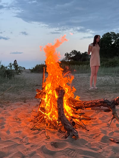 The image depicts an evening scene, likely at the beach in Novo Oriahovo, Bulgaria. A bonfire is prominently burning in the foreground on sand. To the left of the fire, a woman is standing with a drink in her hand. She wears a light-colored dress. Behind the woman and fire, there's some vegetation, possibly dune grass and trees, with the sky visible in the background. The sky has a gradient of colors, indicating either dusk or dawn. There is also a person visible behind the fire.