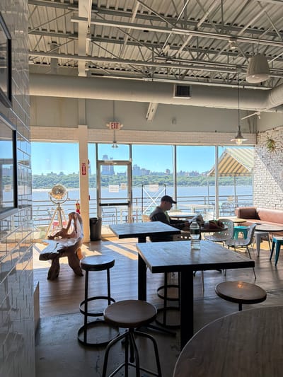 The image depicts the interior of a modern cafe or restaurant with large windows overlooking a body of water, likely the Hudson River, with the Edgewater, New Jersey skyline visible in the distance. The cafe features an industrial-chic aesthetic with exposed ceilings, metal beams, and ductwork.

In the foreground, there are several dark-colored tables and stools. One table in the center has a bottle of water on it. To the left, a large, abstract wooden sculpture sits on the wooden floor.

A man wearing a baseball cap and a dark shirt is seated at a table near the windows, engrossed in reading a newspaper.

The large windows offer a panoramic view of the waterfront, with boats and possibly a ferry visible on the water. Beyond the water, the New Jersey coastline is lined with trees and buildings. The sky is clear and blue, indicating a sunny day.

The cafe is sparsely populated, with only the one man clearly visible as a subject. Other tables and chairs are present, some set up for outdoor seating visible through the windows. The overall ambiance suggests a relaxed and contemporary dining or working space.