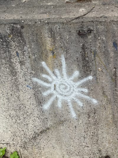 This image captures a close-up view of a rough, grey concrete surface, likely an outdoor setting given the presence of some green foliage in the lower right corner and what appears to be a piece of metal embedded in the concrete on the left side. The concrete itself is textured and speckled with darker and lighter aggregate.
The primary subject is a stylized sunburst symbol spray-painted in white onto the concrete. The symbol has a circular center with concentric rings, from which multiple radiating lines or rays extend outwards. Some of these rays appear more solid than others, and the spray paint is somewhat uneven, giving it a handmade, informal appearance.
To the left of the sunburst, a bent piece of dark metal, possibly a nail or rebar fragment, is protruding from the concrete. The lighting suggests it is daytime, with the sun casting a subtle illumination on the surface. No text is visible in the image. The overall impression is one of an urban or industrial environment, perhaps a wall, pavement, or architectural feature, marked with informal graffiti. The rough texture of the concrete and the presence of weathered elements suggest the location is not new and has been exposed to the elements.