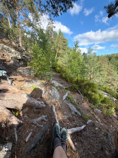 The image depicts a person's legs and feet resting on a forested hillside. The individual is wearing hiking boots and socks, with their legs crossed. The ground is covered in pine needles, rocks, and exposed tree roots. In the background, a dense forest can be seen with a clear blue sky and scattered clouds above. A backpack is visible to the left of the frame, resting on the rocky terrain. The scene appears to be a tranquil outdoor setting, possibly a hiking trail. The location is not identifiable as Ruch'i, Russia based on visual cues.