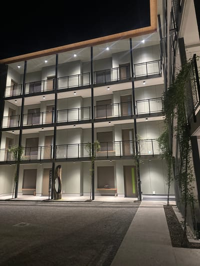 The image shows a multi-story building facade at night, featuring a grid-like structure with balconies or open spaces. The building is pale in color, with dark window coverings. There are plants or vines hanging from the upper levels. A pathway runs alongside the building, with a planter area on the right. A large, hollowed-out log structure serves as a bench or decorative feature near one of the entrances. The location and city of Fehraltorf, Switzerland, cannot be confirmed, as there is no additional context to corroborate this information.