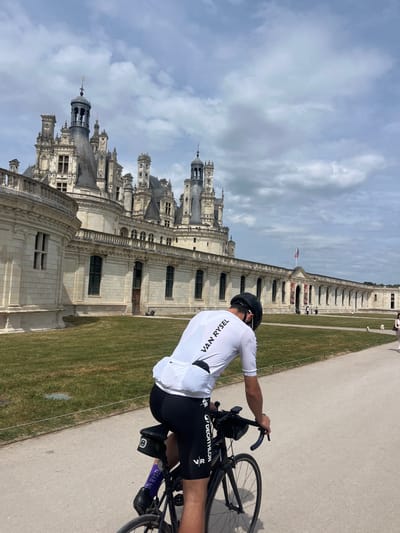 A man is riding a bicycle on a paved path with a grass border alongside a large, ornate building in Chambord, France. The building is made of light-colored stone, with elaborate architectural details including multiple towers, windows, and decorative elements. The sky is partly cloudy. The man is wearing a white cycling jersey with "VAN RYSEL" printed on the back, black cycling shorts with the "VR" logo, and a black helmet. The bicycle appears to be a road bike. The scene is set outdoors, likely in a tourist area near the Château de Chambord.