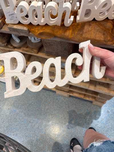 A person's hand, with neatly manicured fingernails, holds a large, three-dimensional white letter that spells out "beach" in cursive. The letter is made of a material that appears to be textured, possibly foam or a similar lightweight substance. The letters are arranged vertically in the image, with the "b" at the bottom and the "h" at the top. The word is being presented against a backdrop of rustic wooden planks and a shimmering golden fabric. In the background, a wooden pallet is visible, and the floor is a mottled grey with faint blue speckles. To the right of the frame, a person's legs are visible, wearing ripped denim shorts and black shoes with pink accents. The scene appears to be indoors, possibly a retail environment given the presence of the pallet and the packaging on the letters. There is no indication of the time of day or weather conditions. The overall mood is casual and perhaps suggestive of a vacation or seaside theme. Visible text on a tag attached to the "b" reads "50H04252S" and a barcode.