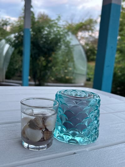 On a white, slatted table, two candle holders are displayed. To the left, a clear glass votive holder contains sand and several seashells. To the right, a turquoise, scaled glass candle holder holds a dark-colored candle. The background is blurred, showing lush green trees and a cloudy sky. The scene is situated on a porch, indicated by the presence of blue and gray wooden posts in the background.