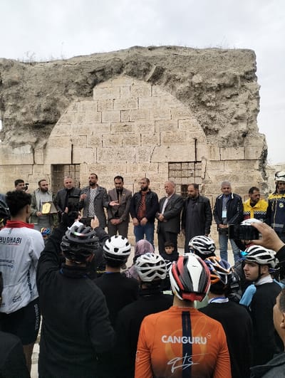 A group of men stand before a large, dilapidated stone structure, possibly an ancient ruin. Several men in business attire are at the forefront, some holding microphones or awards. A crowd of cyclists, identifiable by their helmets and jerseys branded with "ABU DHABI" and "CANGURU," surrounds them. A young boy peeks from behind the men in suits. The presence of emergency personnel suggests an official event or gathering near a historical site.