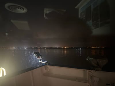 A nighttime view from a boat or ferry. Reflections on the water show a distant city's lights. The boat's interior, visible through the glass, includes a ceiling with circular vents and a window. A spotlight shines on the deck. The overall scene is dark and muted, emphasizing the contrast between the illuminated city and the dark water.