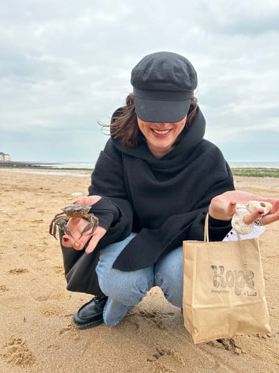 A female individual is depicted crouching on a sandy beach located in Thanet, United Kingdom. The background shows an overcast sky, a calm sea, and a distant shoreline with rocky areas and white cliff-like structures or buildings to the far left. The individual wears a black long-sleeved hooded garment with thumbholes, light blue jeans, and black flat shoes. A black baker boy cap obscures her upper face, revealing a visible smile. In her left hand, which has dark nail polish, she holds a live, dark-colored crab. Her right hand displays multiple light-colored, porous marine objects, resembling coral or weathered shells, with a white lace item visible beneath them. Adjacent to her right leg, a brown paper bag bearing the logo "Rope + Loko Lifestyle Store" rests on the sand. The immediate sand in the foreground shows various impressions.