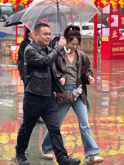 This image depicts an East Asian man and woman walking closely together on a rainy day in Pengshui Miao and Tujia Autonomous County, China. The ground is wet, showing reflections of the overcast sky and vibrant background elements. The man, appearing mid-aged, has short dark hair and wears a black leather jacket, black pants, and dark athletic shoes. He holds a clear umbrella with a white handle, positioned to shield both individuals from precipitation. The woman, appearing younger with dark hair tied in a high ponytail, is smiling. She wears a brown jacket over a plaid v-neck top, light blue flared jeans, and white-soled platform sneakers. A small brown patterned shoulder bag, adorned with a brown ribbon and a grey plush elephant charm, hangs from her shoulder. Her left arm is linked with or adjacent to the man's right arm. In the background, a large red structure with multiple traditional red and yellow lanterns and Chinese characters, including "百业" (bǎiyè), is visible. A white vehicle and another person holding a black umbrella are partially obscured in the distance.