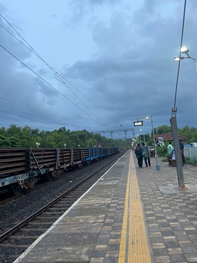 The image depicts a train station platform in Sircaim, India, with a moody, overcast sky. The platform itself is paved with dark and light grey square tiles, bordered by a textured yellow tactile paving strip. The train tracks run alongside the platform, with a freight train consisting of multiple carriages visible. The train carriages appear to be carrying some sort of metal components or materials.

Several people are present on the platform. In the foreground, two individuals stand with backpacks, appearing to be waiting. Further down the platform, more people are visible, some sitting and others standing. One person wears a light green shirt, and another has a dark blue shirt. A sign with the number "18" is mounted on a pole.

Overhead, a streetlamp with two lights illuminates the scene, casting a warm glow against the dull sky. Electrical wires crisscross the sky, supported by poles. A digital clock displaying "01:20" is mounted on a pole above the platform. The background features lush green trees, suggesting the station is surrounded by nature. The overall atmosphere is somewhat somber due to the heavy clouds, but the lights and the presence of people indicate ongoing activity at the station. There are no prominent signs of distress or urgency, suggesting a normal day at the station.