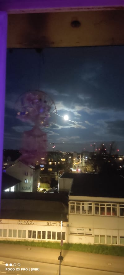 The image is an eye-level nighttime shot of a street scene in Constance, Germany. The buildings are situated along a street. The building on the right has multiple windows and a sign that reads "velotours". Adjacent to it, graffiti is present with partially visible text saying "FAHR SCHU...P. 92.XX. GOT.2". Overhead are clouds lit by what appears to be moonlight. In the foreground, there's a spherical object, possibly a decoration. The photo may have been taken from inside a building looking outward. There is a smartphone's watermark "POCO X3 PRO | [WIR]" in the bottom right corner.