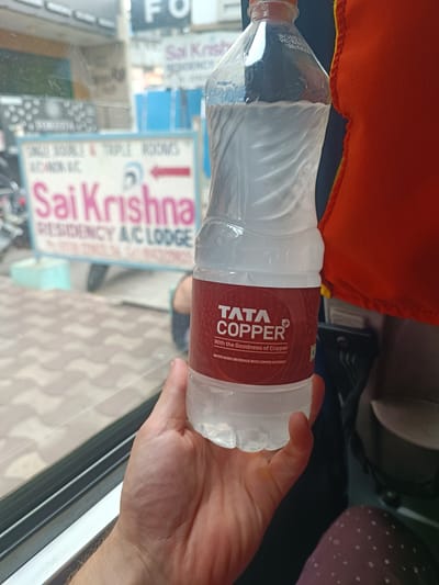 A person's hand is holding a plastic bottle of Tata Copper water. The bottle has a red label with white text that reads "TATA COPPER" and "With the Goodness of Copper". The bottle is mostly filled with clear liquid. In the background, a sign for "Sai Krishna RESIDENCY A/C LODGE" is visible. This sign suggests the location is in Kurnool, India, as Sai Krishna Residency is a known hotel in Kurnool. The environment appears to be indoors, possibly a vehicle, with a window showing a blurred outdoor scene.