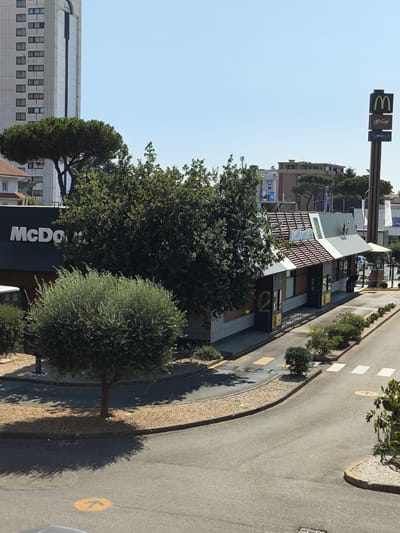 This image depicts a bright, sunny day at a McDonald's restaurant with a McCafe section in Pomezia, Italy. The McDonald's building has a distinctive roof design with red and white striped elements. Several drive-thru windows are visible, labeled with "PAGA" (Pay) and "RITIRA" (Collect). A large, dark green McDonald's sign with the iconic golden arches and "McCafe" branding stands tall on the right side of the frame.
In the foreground, a small, rounded tree with lush green foliage is positioned in front of the McDonald's. The drive-thru lane curves around this tree, with yellow markings and white crosswalks on the asphalt. Gravel and small green bushes line the edges of the drive-thru lane and surrounding areas.
In the background, a tall, modern white building with numerous windows dominates the skyline. Other buildings with varying architectural styles are also visible, along with mature trees. The sky is clear and blue. The overall scene is clean and well-maintained, suggesting a typical commercial area. There are no people or vehicles clearly identifiable in the image.