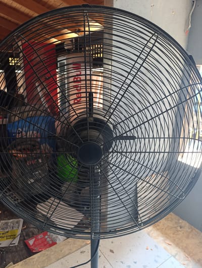 This media file depicts a large, black, industrial-style oscillating fan positioned in an indoor, likely workshop or storage area. The fan is the primary subject, taking up most of the frame with its intricate wire grille. Behind the fan, various objects are partially visible, including what appears to be a blue container or machine, a green bucket, and some printed materials on a wooden surface, one of which has text that is partially legible as "Abdeck-Plak". The background suggests a utilitarian setting with wooden beams on the ceiling and a wall that is partially tiled. The lighting is bright, indicating it is daytime. There is no activity taking place other than the fan being present. The overall impression is of a functional, perhaps somewhat cluttered, workspace.