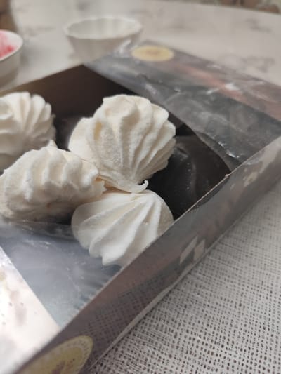 The image shows a close-up of a box of white marshmallows, likely zephyr, on a textured white tablecloth. The marshmallows are fluffy and have a distinctive swirl pattern. One marshmallow is slightly out of focus in the background, while three are in sharp focus in the foreground. The box is clear plastic, revealing the marshmallows inside. The packaging has some text and graphics, including a yellow circular logo. In the blurred background, there are two small white bowls, one appearing to contain a pink substance. The lighting suggests it's daytime, and the setting appears to be indoors, possibly a home environment. The overall mood is casual and domestic, with a focus on the sweet treat. No people are visible in the image.