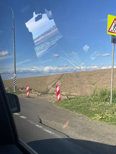 The image shows a roadside scene with a dirt road and a pile of earth in the background under a blue sky with clouds. A pedestrian crossing sign is visible on the right. There are two red and white striped traffic barriers on the road. A light pole with black and white diagonal stripes is on the left. The image appears to be taken from inside a vehicle, as suggested by the reflection on the windshield and the presence of a side mirror on the left. The location is identified as Starye Chernitsy, Russia.