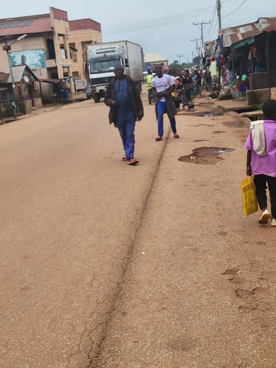 In Tangu, Nigeria, a street scene unfolds under an overcast sky. A man in a dark jacket and blue trousers walks down the center of a dusty, cracked asphalt road. Behind him, a younger man in a white t-shirt and blue trousers, smiling, walks with a jacket slung over his shoulder. Further down the road, a person in a purple t-shirt and black trousers walks away from the camera, carrying a yellow bag. On the right side of the road, stalls with corrugated metal roofs display colorful wares, with chairs stacked nearby. A large white truck with "UB NATIONAL" written on it is parked on the left side of the road. Buildings line the left side of the street, some featuring advertisements. Power lines stretch across the sky, and a street lamp is visible on the left. The ground is littered with puddles and debris, indicating recent or ongoing rain.