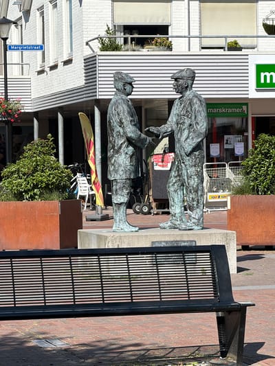 This is a vertical, eye-level outdoor shot of a bronze statue depicting two men in historical clothing. The statue is situated on a paved area with a red brick ground. To the right of the statue, a black metal bench with a slatted seat is visible. In the background, there are buildings with a corrugated metal facade and a white brick facade. A green sign with a white "m" is attached to the corrugated facade, and below it, text reads "marskramer." A bicycle with a yellow and red flag is leaning against a metal post near the statue. The scene appears to be in a town square or a commercial area. The lighting suggests it is daytime, possibly sunny with shadows cast on the ground. The overall impression is a public art installation in a Dutch town. A street sign visible in the lower left corner reads "Margrietstraat."
