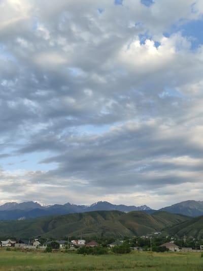 The image depicts a rural landscape in Dolan, Kazakhstan, under a dramatic, cloudy sky. The foreground is dominated by a vibrant green field of grass, suggesting a natural, undeveloped area. Behind the field, a cluster of houses forms a small village, characterized by a variety of roof colors, including brown, white, and dark grey. The houses appear to be single-story or two-story residential buildings with surrounding fences and some greenery.

Beyond the village, rolling green hills rise, their slopes textured with subtle contours and shadows that indicate uneven terrain. The hills are covered in lush vegetation, contributing to the overall pastoral feel of the scene. In the distance, majestic mountains form a formidable backdrop. Some of these peaks are capped with snow, even in what appears to be a warmer season, adding a sense of grandeur and ruggedness.

The sky is a prominent feature, filled with a complex arrangement of clouds. Large, billowing grey clouds dominate, interspersed with patches of bright blue sky. Sunlight filters through some of the clouds, creating areas of luminescence and dappled light on the landscape below. The interplay of light and shadow across the clouds suggests a dynamic and possibly changing weather pattern.

The overall mood of the image is one of tranquility and natural beauty, with a hint of the wildness of the distant mountains. The time of day appears to be late afternoon or early evening, given the soft lighting and the positioning of the sun, which is not directly visible but inferred from the illumination. There are no visible people or specific objects of note beyond the natural elements and the village structures. No text is present in the image.