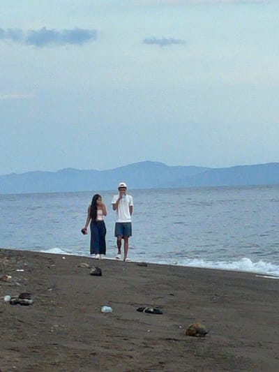 The image depicts a beach scene in Dauin, Philippines, with two individuals walking near the water's edge. The ground is dark sand, scattered with debris including plastic bottles and what appear to be flip-flops. The ocean is calm with gentle waves breaking on the shore. In the background, a mountain range is visible under a hazy, overcast sky. One person, a female, is wearing a strapless top and a long, dark skirt. The other person, a male, is wearing a white t-shirt, dark shorts, and a white cap. He is holding a light-colored object, possibly a phone, near his face. The overall lighting suggests it is either early morning or late afternoon.