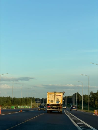 The image depicts a highway scene with a truck in the foreground. The truck is white and appears to be carrying some cargo, with branding that includes "MKM3 LIGHTS" and "4200 KILOGRAMS". It has a license plate that reads "BE 6704 AM". To the left of the truck, a black car is visible, with a license plate that appears to be "X 084 CA 154". Further down the road, a blue SUV is visible, with a license plate that is not clearly legible. The highway is paved and has a white dashed line marking. On the right side of the road, there are traffic cones and construction barriers. A line of streetlights stretches along the side of the road, and beyond that, a dense line of green trees forms the background. The sky is a clear blue with some wispy clouds. The overall impression is that of a typical highway scene during daylight, likely in a rural or suburban area given the surrounding trees. There is no indication of the specific location within the city of Kaipy, Russia, as no landmarks or specific geographical features are identifiable.