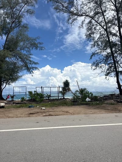 The image depicts a roadside view of a beach area in Ban Tha Chat Chai, Thailand, on a partly cloudy day. In the foreground, a gray asphalt road with a white line runs horizontally across the bottom of the frame. Behind the road, a strip of dirt and vegetation separates it from the beach.
The beach itself features scattered debris, including what appears to be a blue plastic container and some white debris, possibly Styrofoam. There are several upright metal frames, possibly remnants of a fence or some form of structure, positioned on the sandy area. A swing set with two seats is visible to the left, with one seat occupied by a person wearing a pink top and dark bottoms.
The ocean is visible beyond the beach, with moderate waves. The sky is a vibrant blue, punctuated by large, puffy white cumulus clouds. Two large trees, characterized by their tall, thin trunks and sparse, needle-like foliage, frame the scene on the left and right sides, their branches reaching towards the sky. Lush green vegetation, including palm-like plants, grows on the beach area. In the distance, the ocean meets the horizon under the expansive sky.