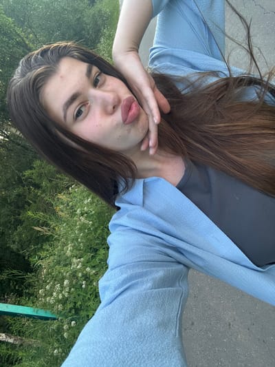 This is a selfie of a young woman in an outdoor setting, likely in Шевлягино, Russia. The image is oriented sideways, with the woman's head towards the bottom of the frame. She has long, brown hair and is wearing a dark grey t-shirt and a light blue, textured open cardigan or jacket. Her left hand is raised to her face, partially covering her mouth, and her lips are slightly parted, giving her a pouting or playful expression. Her eyes are looking towards the camera with a neutral to slightly curious gaze.

The background consists of lush green foliage, suggesting a natural environment with trees and plants. There are some small white flowers visible among the greenery, and a portion of a light grey paved surface, possibly a sidewalk or path, is also in view. A thin, bright green vertical object, possibly a post or railing, is visible on the right side of the frame. The lighting suggests it is daytime, and the outdoor setting with green vegetation indicates pleasant weather. The overall mood of the image is casual and relaxed. There is no visible text in the media file.