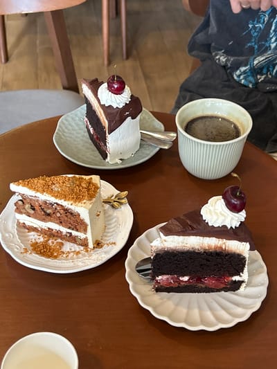 This image captures a close-up, overhead view of a wooden table in a cafe located in Chiang Dao, Thailand, laden with three slices of cake and two beverages. In the top right corner, the upper torso of a person is partially visible, wearing a dark garment with light blue patterns, and a hand with a ring on the finger is present.

From left to right and top to bottom:
A slice of carrot cake rests on a white, scalloped-edge plate in the bottom left. The cake features multiple layers of orange-hued cake interspersed with nuts and white cream cheese frosting, topped with brown crumbs. A small, gold-colored leaf-shaped cake fork is inserted into the cake.
Centrally placed on the table is a slice of layered chocolate cake on a light green, textured plate. This cake has dark chocolate cake layers, a white cream layer, a lighter brown cream layer, and is coated with dark chocolate ganache. It is garnished with a dollop of white whipped cream and a single red cherry with a green stem. A silver spoon lies to its right.
To the right of the carrot cake, a white ceramic cup, partially visible in the bottom left, contains a light, milky liquid.
Above the bottom right slice of cake, a light green, ribbed ceramic mug contains dark black coffee.
In the bottom right, another slice of chocolate cake is presented on a white, scalloped-edge plate. This cake features a dark chocolate cake base, a distinct layer of red fruit filling (likely cherry or berry compote), topped by a thick layer of white whipped cream and dark chocolate ganache. It is also garnished with a dollop of white whipped cream and a single red cherry with a green stem. A silver fork is positioned next to it.
The table is made of smooth, reddish-brown wood. In the blurred background, parts of light brown wooden furniture legs and light grey upholstered seating are visible, indicating an indoor cafe environment.