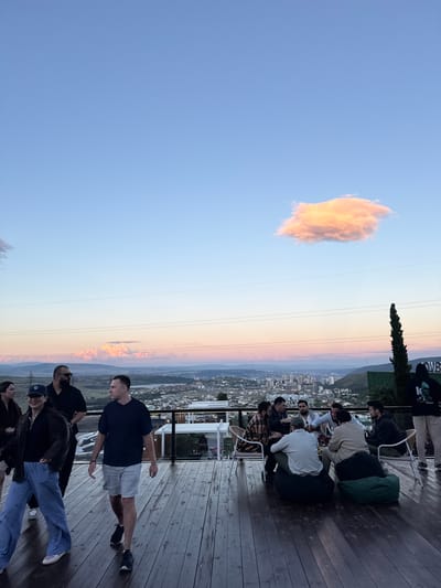 A group of people are gathered on a wooden deck overlooking a cityscape and distant mountains at dusk. In the foreground, two individuals are walking on the deck. A woman in striped pants and a dark jacket is walking towards the left of the frame, and a man in a dark t-shirt and light shorts is walking towards the right. Several other people are seated around a table and on beanbag chairs in the middle ground, engaged in conversation. A tall, thin tree stands to the right of the group, and a sign with the word "LOVE" is partially visible on a wall behind them. The sky above is a gradient of soft blues and pinks, with a single illuminated cloud. The cityscape extends into the distance, with buildings nestled among rolling hills. The overall scene suggests a relaxed social gathering at an outdoor venue.