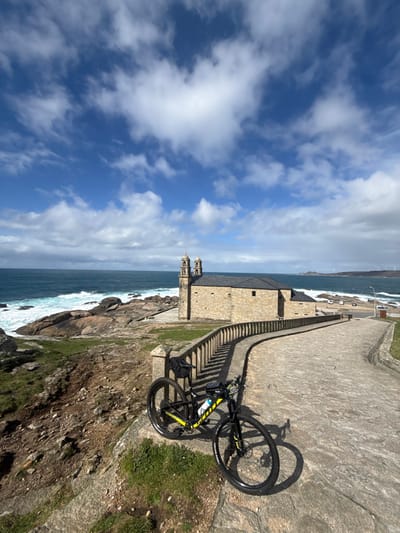 The scene depicts a coastal landscape in Muxía, Spain, centered around the Sanctuary of Nosa Señora da Barca. In the immediate foreground, a black and yellow full-suspension mountain bike is parked, leaning against a low, curved stone balustrade that outlines a paved stone pathway. The pathway extends from the lower right towards the middle ground. To the left of the path and balustrade, the terrain drops off into a rocky, grass-covered cliffside leading to the ocean.

In the midground, the Sanctuary of Nosa Señora da Barca, a historic stone church featuring two prominent bell towers and a dark grey roof, stands on a rocky outcrop directly adjacent to the Atlantic Ocean. The ocean is characterized by turquoise waters with white-capped waves crashing against the rugged, dark grey rock formations along the coastline.

The background features the open ocean extending to the horizon under a bright blue sky with numerous scattered white cumulus clouds. To the far right, a distant headland or coastline is visible, marked by several wind turbines. No individuals are present in the image.