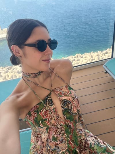 A woman wearing sunglasses and a paisley-patterned dress poses outdoors, likely on a cruise ship deck with a view of the ocean. She faces slightly right with her head tilted to the left. The deck features wooden planks, and a glimpse of a turquoise chair is visible. The ocean stretches to the horizon, with a rocky coastline visible in the distance.