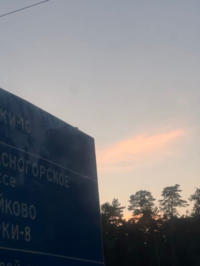 A dark blue road sign with white text is angled from the bottom left towards the center of the frame. The text is in Russian and partially obscured by the angle and focus. Visible text includes "КИ-10", "CHOGORSKOE", "ce", "IKOVO", and "КИ-8". The sign is set against a pale sky with a faint orange cloud streak near the horizon. Silhouetted trees line the horizon beneath the sky. A thin dark line, possibly a power cable, runs diagonally across the upper left portion of the sky. The overall scene depicts a road sign and a natural landscape at dusk. The location is identified as Usovo, Russia.