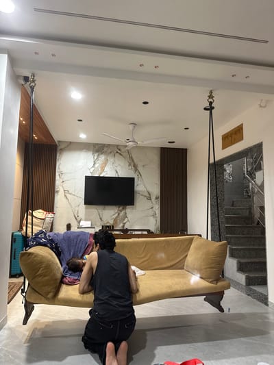 The image displays an indoor residential scene in Surat, India. An adult individual is observed from behind, kneeling on a light-colored tiled floor, facing a mustard-colored swing sofa (jhoola). The individual wears a dark sleeveless top and dark pants/shorts. An infant lies on the left side of the swing sofa, partially covered by blue and patterned fabric.
The swing sofa is suspended from a white false ceiling by black ropes and brass-toned fixtures. In the background, a large flat-screen television is mounted on a wall featuring white marble with grey veining. This marble section is flanked by vertical dark wooden slats. A white ceiling fan and multiple recessed lights are visible in the ceiling. To the left, a light-colored wheeled suitcase and a fabric bag are present on the floor. To the right, an open doorway reveals a multi-tiered staircase with dark treads and a metal handrail. A wooden plaque with script is mounted on the wall above the doorway. A dark brown wooden structure resembling a console table or railing is visible behind the swing sofa. Several small red objects are on the floor in the bottom right corner of the frame.