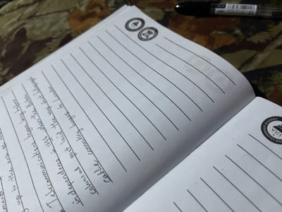 The image shows an open notebook with lined pages. There are two circular emblems at the top of the left page, each with a design inside. Handwritten text is visible on the left page, starting with "The British" and continuing with "The telecommunications" and "in dependence" followed by "cable". A black pen is positioned horizontally in the upper right corner of the image, resting on a patterned surface that appears to be fabric, possibly a camouflage-patterned cloth. On the right page, a circular emblem with the words "LILA LEADERSHIP ACADEMY" is visible. The notebook is placed on the patterned fabric surface. There is no indication of the city of Alasia, Nigeria, or any specific urban environment.