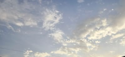 The image shows a cloudy sky over Margilan City, Uzbekistan. Thin, wispy clouds are scattered across a pale blue sky, with some denser, more textured clouds present, particularly towards the right side of the frame where they are illuminated by sunlight. A thin, dark horizontal line, possibly a power line, is visible in the lower portion of the image. No people, animals, or structures are depicted, focusing solely on the atmospheric conditions.