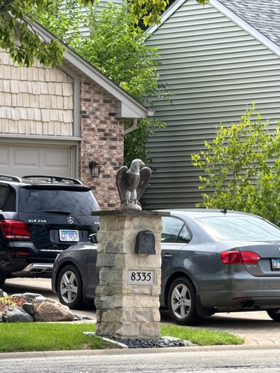 This is a vertical, eye-level, outdoor shot of a residential driveway in Woodridge, United States of America. The scene captures a stone pillar with a mailbox and the number "335" displayed on it. A decorative eagle statue is perched atop the pillar. Two cars are visible: a dark colored SUV parked behind the pillar and a dark grey sedan parked further down the driveway. The houses in the background feature light-colored siding and brick accents. Lush green trees and a well-maintained lawn add to the suburban setting. The time of day appears to be daytime, with bright natural lighting, and the weather seems fair. No people are present in the image. The visible text includes the number "335" and a license plate on the SUV reads "Z31 0263". The sedan's license plate is partially visible and shows "CJ" and "JETTA" is visible on the rear of the sedan.