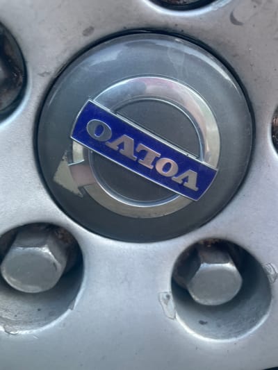 The image is a close-up, eye-level shot of a car's wheel. The focus is on the center cap, which features a blue rectangular badge with the silver "VOLVO" logo. The wheel itself is silver-colored and appears to be made of alloy, with visible lug nuts. The surrounding environment is out of focus. The image was taken in Бабовіцкі сельскі Савет, Belarus.